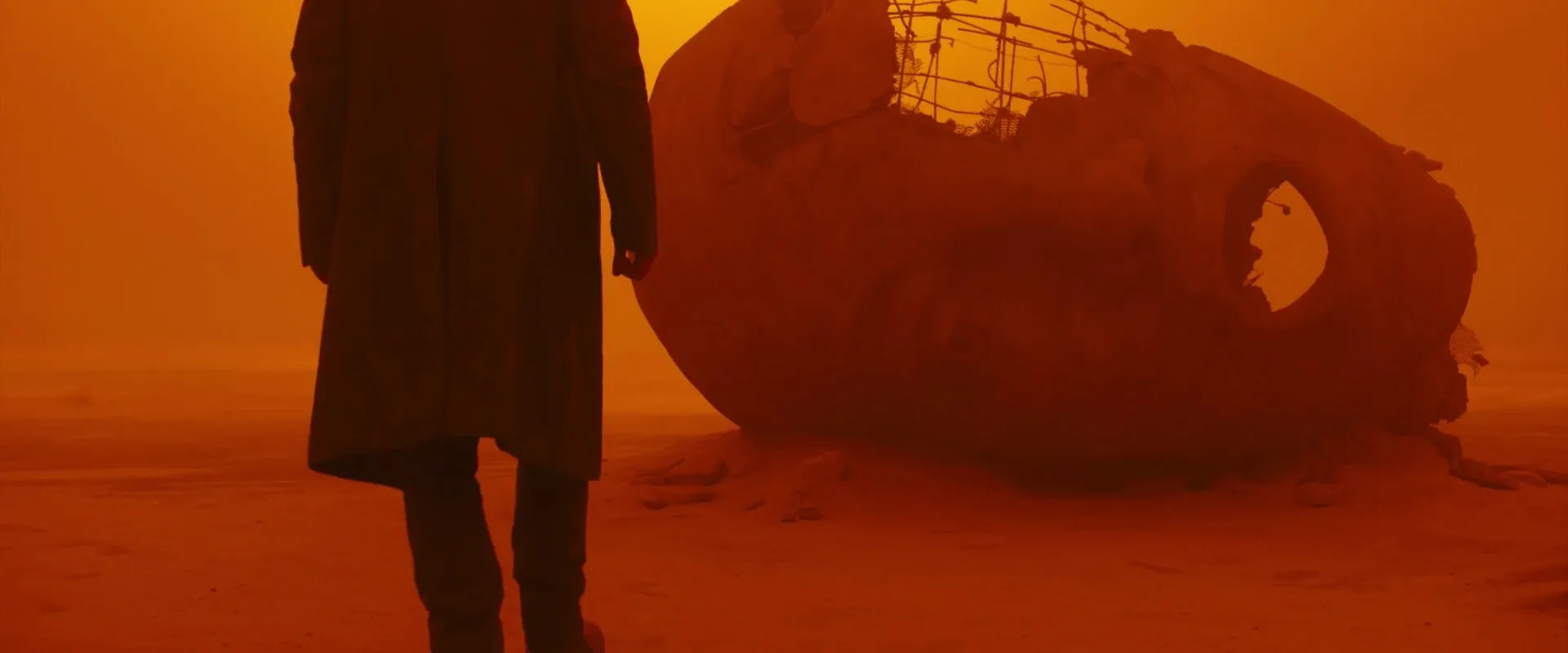 Blade Runner 2049 (2017) β K stands beside a toppled statue's head, dwarfed by the orange haze of the wasteland