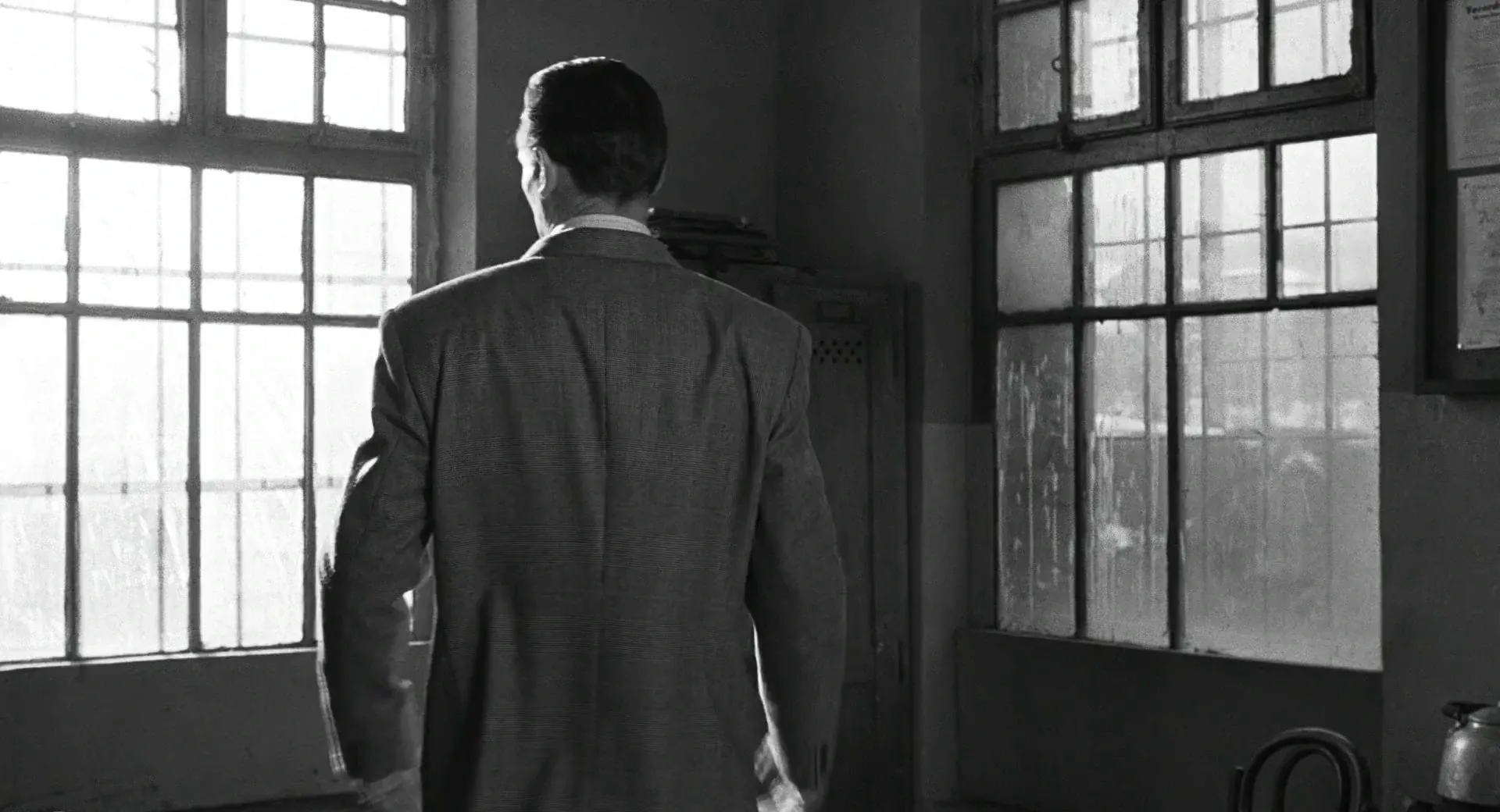 Janusz Kamiński's black-and-white cinematography for Schindler's List (1993): a man seen from behind, held at a distance, backlit by tall barred windows