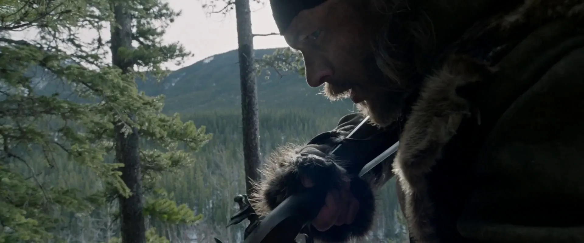 A weathered frontiersman in profile in The Revenant, lit only by soft overcast daylight against a forested mountain slope