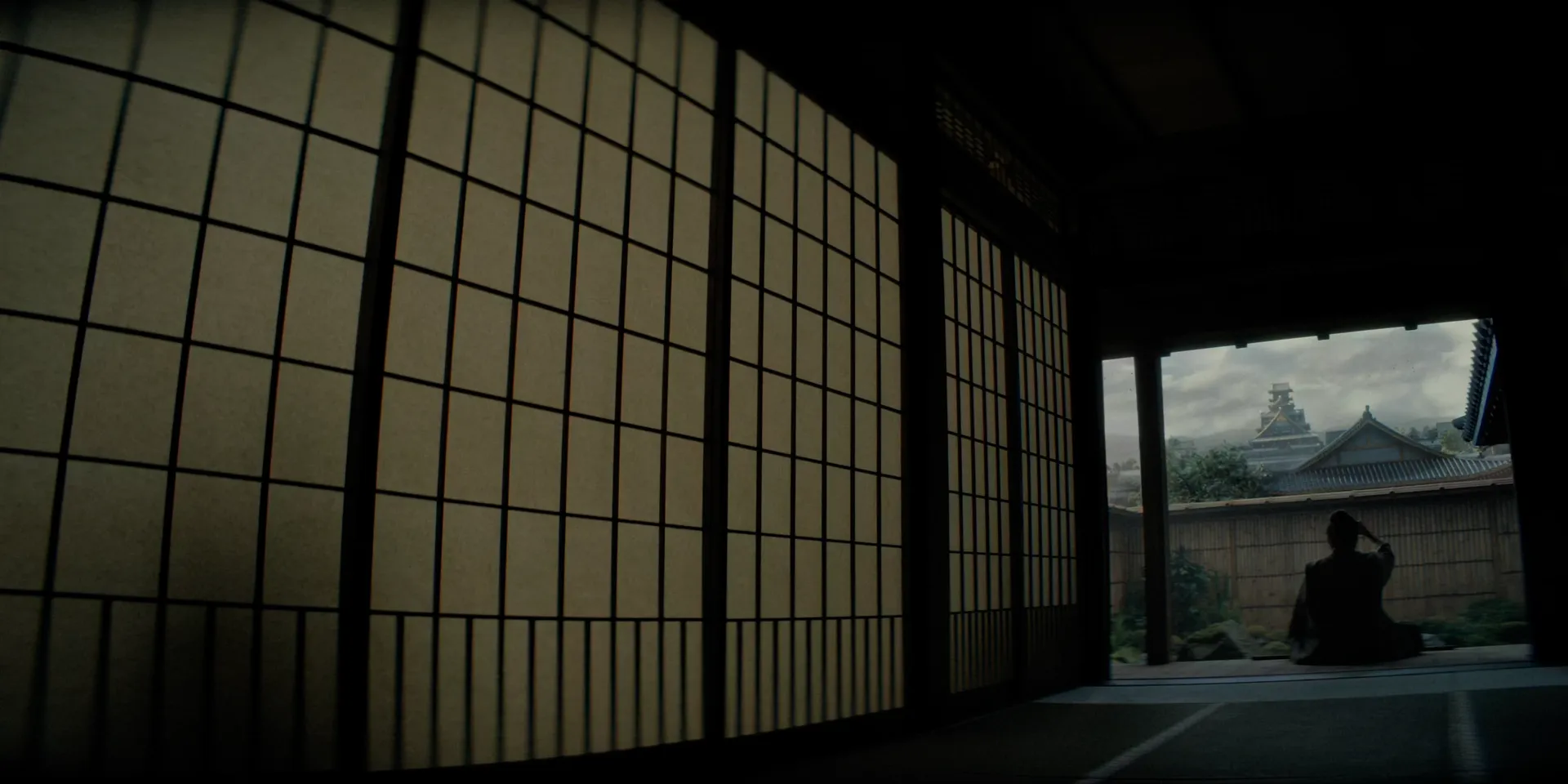 A still, locked-off frame: shoji screens fill the left of the composition while a lone figure sits framed against a castle beyond the veranda β Shogun (2024)
